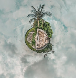 A tiny planet-style image showcases a small sphere covered with greenery and a single palm tree at the top. In the center, there is a rooftop and part of a building, surrounded by lush vegetation. The sky forms a circular background with a mix of cloudy and clear patches.