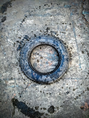 A single black tire lies flat on a rough, textured concrete surface. Water stains and dark patches surround the tire, indicating moisture or rain. The tire shows wear and tear, suggesting it might be old or used.