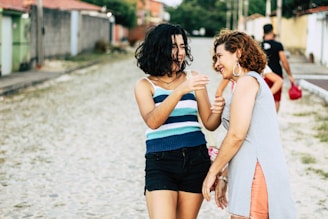 Two women are walking and laughing together on a cobblestone street, surrounded by colorful buildings and greenery. They appear relaxed and engaged in conversation, sharing a joyful moment.