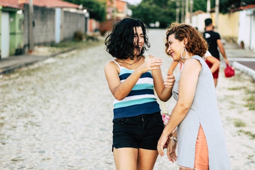 Two women are walking and laughing together on a cobblestone street, surrounded by colorful buildings and greenery. They appear relaxed and engaged in conversation, sharing a joyful moment.