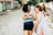 Two women are walking and laughing together on a cobblestone street, surrounded by colorful buildings and greenery. They appear relaxed and engaged in conversation, sharing a joyful moment.