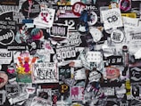 A densely packed collage of various stickers, posters, and graffiti featuring a diverse mix of designs and text in different colors and styles. The overall theme is chaotic and colorful, showcasing elements of urban street art and popular culture.