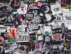 A densely packed collage of various stickers, posters, and graffiti featuring a diverse mix of designs and text in different colors and styles. The overall theme is chaotic and colorful, showcasing elements of urban street art and popular culture.