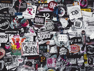 A densely packed collage of various stickers, posters, and graffiti featuring a diverse mix of designs and text in different colors and styles. The overall theme is chaotic and colorful, showcasing elements of urban street art and popular culture.