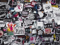 A densely packed collage of various stickers, posters, and graffiti featuring a diverse mix of designs and text in different colors and styles. The overall theme is chaotic and colorful, showcasing elements of urban street art and popular culture.