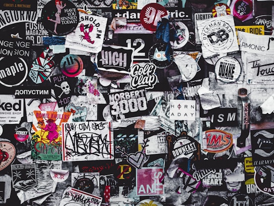 A densely packed collage of various stickers, posters, and graffiti featuring a diverse mix of designs and text in different colors and styles. The overall theme is chaotic and colorful, showcasing elements of urban street art and popular culture.