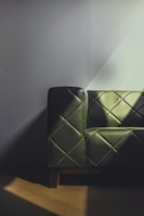 A leather sofa with dark green upholstery featuring a quilted pattern. The design includes diagonal stitching that forms diamond shapes. Soft light casts shadows across the sofa and wall, creating a mix of light and dark areas.