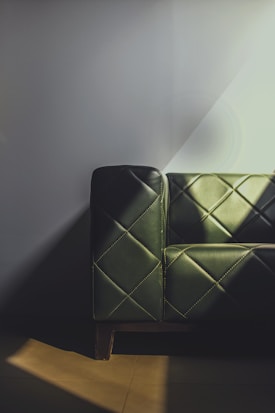 A leather sofa with dark green upholstery featuring a quilted pattern. The design includes diagonal stitching that forms diamond shapes. Soft light casts shadows across the sofa and wall, creating a mix of light and dark areas.