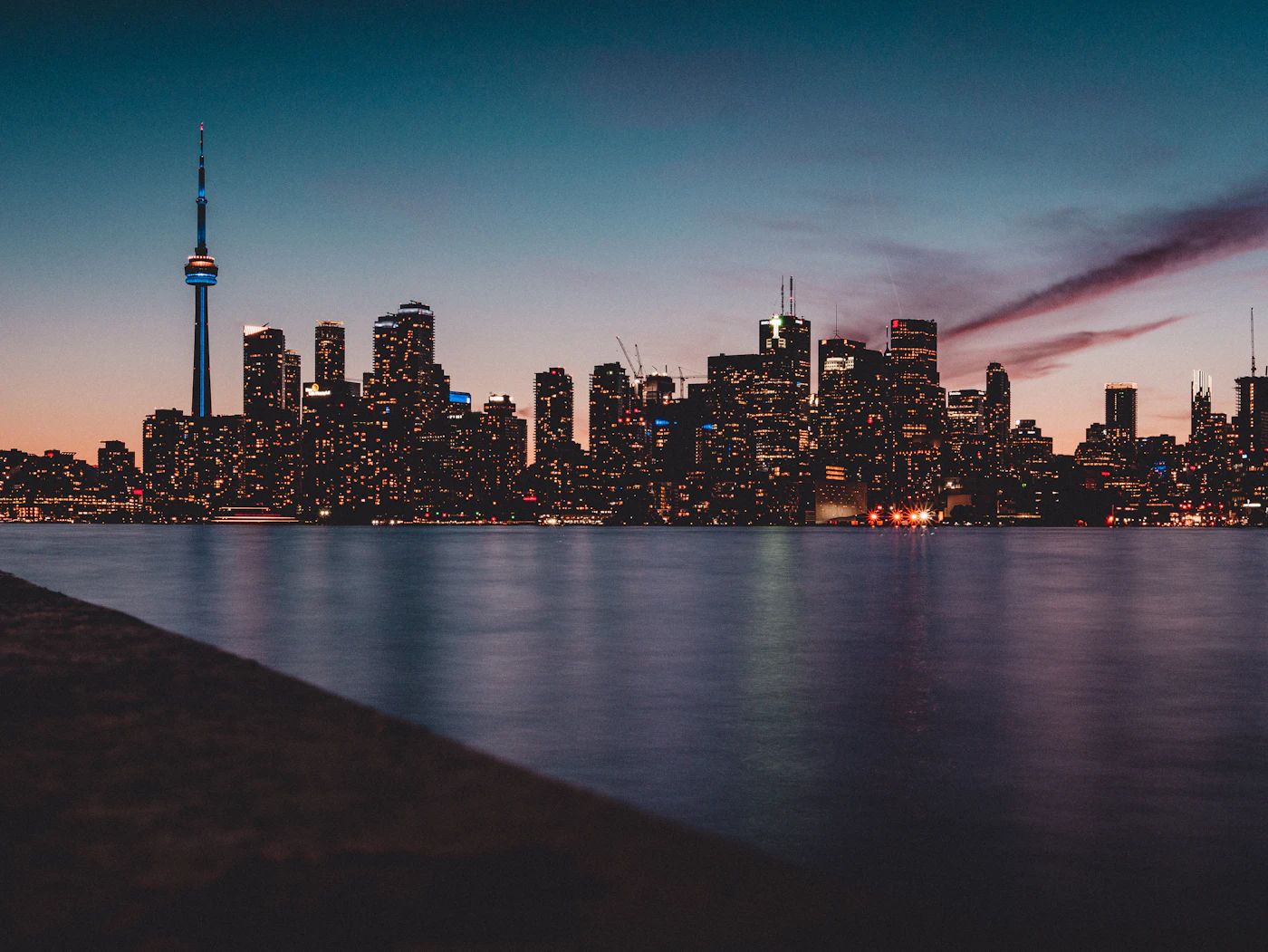 The Toronto skyline glowing at dusk across calm water, the CN Tower rising above the downtown towers