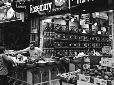 A busy market scene with jars lined up on shelves behind a vendor. The vendor is interacting with a customer, possibly selling a variety of goods placed in large bowls at the stall. Signs in the background display the word 'Rosemary' and other text in different languages.