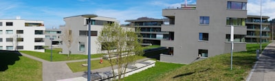 Modern apartment buildings with multiple stories are situated in a residential area. The buildings are surrounded by neatly cut grass and young trees. A few street lamps and paved pathways run through the area, with clear skies above providing a bright and open atmosphere.
