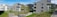 Modern apartment buildings with multiple stories are situated in a residential area. The buildings are surrounded by neatly cut grass and young trees. A few street lamps and paved pathways run through the area, with clear skies above providing a bright and open atmosphere.