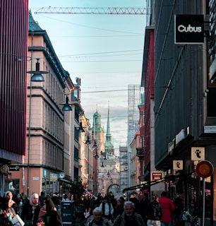 A bustling city street lined with historical buildings and modern shops. Crowds of people are walking along the street, with a prominent church or cathedral visible in the background. Overhead, a construction crane indicates ongoing development in the area. Streetlights and shop signs add to the urban atmosphere.