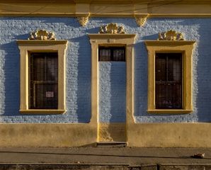 A building facade painted in light blue with yellow trim. The wall features two windows and a door, each framed with decorative elements in a classic style. The windows have black wrought iron grilles, and the surface appears to be made of textured brick. Sunlight casts shadows on the wall, highlighting the architectural details.
