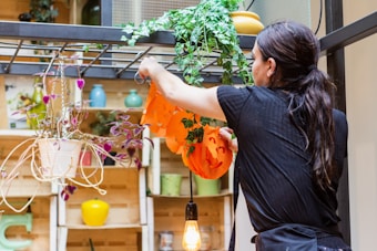 A person is decorating an indoor area with Halloween-themed items, specifically hanging orange pumpkin decorations. Surrounding the area are various indoor plants, some hanging in pots. Shelves display vibrant decor pieces like small vases in different colors. The setting is well-lit with hanging light bulbs.