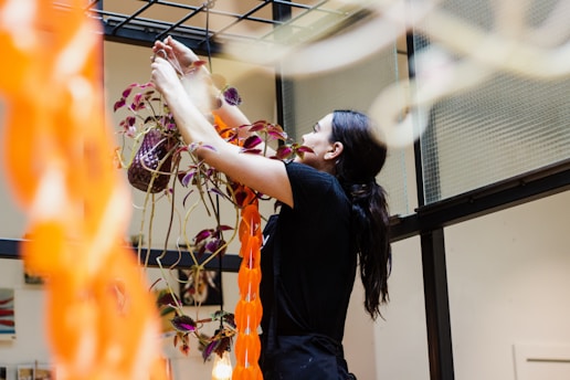 A person with long dark hair is arranging a potted plant with trailing vines and purple leaves. The setting appears to be indoors, with a modern architectural backdrop featuring glass panels and a metal frame. An orange decorative element hangs in the foreground, partially obscuring the view.