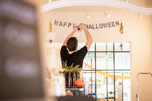 A person is decorating an indoor space for Halloween. They are hanging a banner that says 'Happy Halloween' with fake spiders and web decorations around it. The area is well-lit, and various bottles and a pumpkin can be seen on a table in the foreground. The background features a large window with more decorations.
