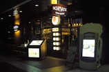 A dimly lit entrance to a restaurant named Schmatz Beer Dining, featuring illuminated signs and menus outside. The interior lights create a cozy ambiance, with people visible inside. The exterior includes a large menu board shaped like a beer glass and a smaller board listing menu items along with lighting elements that enhance the inviting atmosphere.