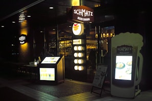 A dimly lit entrance to a restaurant named Schmatz Beer Dining, featuring illuminated signs and menus outside. The interior lights create a cozy ambiance, with people visible inside. The exterior includes a large menu board shaped like a beer glass and a smaller board listing menu items along with lighting elements that enhance the inviting atmosphere.