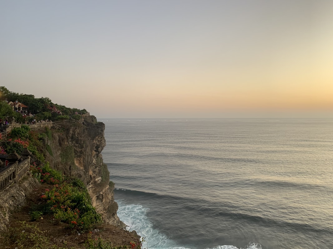 A dramatic cliff edge rises from the ocean, with waves crashing against the rocks below and blue water extending to the horizon under a clear sky.. Photo by Sage Atlas on Unsplash.