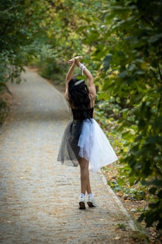 A young girl in a black and white tutu dress is walking down a narrow, tree-lined path. Her arms are gracefully raised above her head, and she appears to be dancing. The path is covered in fallen leaves, and the surrounding greenery creates a serene, natural atmosphere.