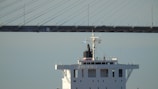 The image features the upper structure of a large ship with a bridge in the background. The ship's deck is visible, along with several antennas and flags. The sky is clear and blue, and the bridge has a modern, cable-stayed design.
