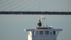 The image features the upper structure of a large ship with a bridge in the background. The ship's deck is visible, along with several antennas and flags. The sky is clear and blue, and the bridge has a modern, cable-stayed design.