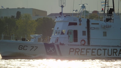 A maritime patrol vessel with the identification 'GC 77' is navigating through waters. The ship's hull is marked with the word 'PREFECTURA' and an anchor symbol. The vessel is set against a backdrop of greenery and urban architecture, with flags visible near the top.