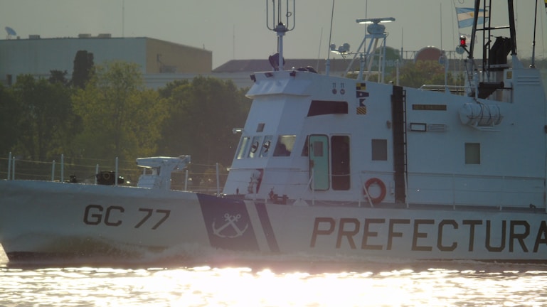 A maritime patrol vessel with the identification 'GC 77' is navigating through waters. The ship's hull is marked with the word 'PREFECTURA' and an anchor symbol. The vessel is set against a backdrop of greenery and urban architecture, with flags visible near the top.
