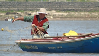 A person is sitting in a small blue fishing boat on a body of water, pulling in a fishing net. The individual is wearing a red vest over a green shirt and a wide-brimmed hat. In the background, there is a blurred view of the shore.