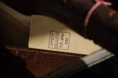 A close-up view of a parcel or packaging with a label containing text in Hebrew and English. The label reads 'JERUSALEM P.P. 9258' and is attached to what looks like a rolled-up item secured with a string. The background shows various shades of brown materials, suggesting a rustic or vintage setting.