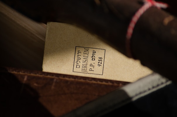 A close-up view of a parcel or packaging with a label containing text in Hebrew and English. The label reads 'JERUSALEM P.P. 9258' and is attached to what looks like a rolled-up item secured with a string. The background shows various shades of brown materials, suggesting a rustic or vintage setting.