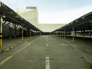 An empty parking lot with rows of covered parking spaces on both sides, featuring yellow and black striped poles. The concrete ground has faded white lines and some patches of grass. A large, plain building is in the background under a partly cloudy sky.