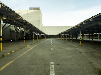 An empty parking lot with rows of covered parking spaces on both sides, featuring yellow and black striped poles. The concrete ground has faded white lines and some patches of grass. A large, plain building is in the background under a partly cloudy sky.