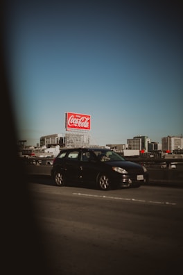 A black car is driving on a highway with a large red Coca-Cola billboard in the background. Modern apartment buildings are visible under a clear blue sky, creating an urban setting.
