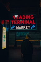 A person wearing a cap is standing near a lamppost at night. Bright neon lights display the words 'Reading Terminal Market' above the scene. There is also a sign advertising the lottery. The setting suggests a quiet and slightly mysterious urban context.