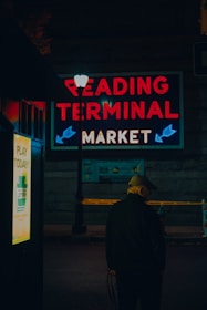 A person wearing a cap is standing near a lamppost at night. Bright neon lights display the words 'Reading Terminal Market' above the scene. There is also a sign advertising the lottery. The setting suggests a quiet and slightly mysterious urban context.