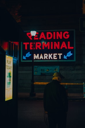 A person wearing a cap is standing near a lamppost at night. Bright neon lights display the words 'Reading Terminal Market' above the scene. There is also a sign advertising the lottery. The setting suggests a quiet and slightly mysterious urban context.
