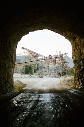 A view from inside a dark tunnel looking out toward an industrial quarry setting. Large, rusted metal structures and conveyors are visible outside, surrounded by piles of gravel and rocky terrain. The atmosphere is rugged, with natural and industrial elements contrasting sharply.