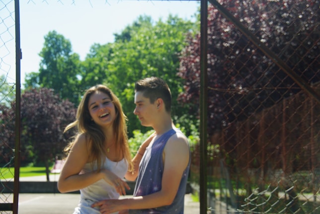A young couple stands outdoors near a mesh fence, surrounded by lush green trees and bright sunlight. The girl is laughing and the boy appears to be in conversation with her. The setting suggests a park or a recreational area.