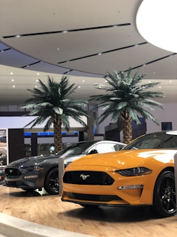 A showroom setting with two Mustang cars prominently displayed. The car on the right is orange, while the one on the left is dark gray. They are positioned on a wooden platform with palm trees in the background. The ceiling is modern and stylish, featuring recessed lighting.