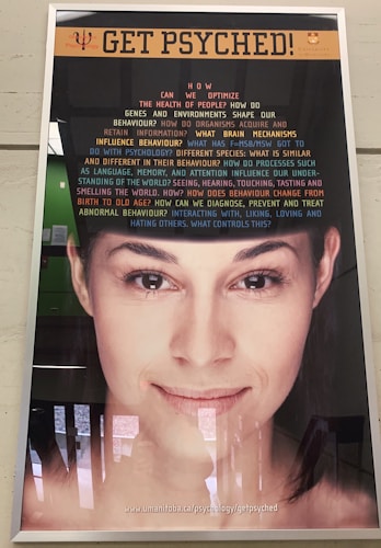 A poster features a close-up of a woman's face with a message about psychology. The top of the poster has the phrase 'GET PSYCHED!' in large orange text. Below, there is a series of questions in varying sizes and colors, exploring different psychological topics. The bottom part of the poster contains a web address.