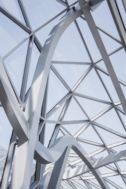 A geometric ceiling design featuring triangular glass panels supported by a network of white, curved metal beams. Natural light floods through the transparent sections, creating an airy and modern architectural appearance.