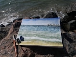 A watercolor painting lies on a rocky shore, depicting a seascape with blue and green hues, small waves, and a city skyline in the distance. The painting is surrounded by art supplies, including brushes and small paint pots.