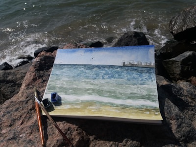 A watercolor painting lies on a rocky shore, depicting a seascape with blue and green hues, small waves, and a city skyline in the distance. The painting is surrounded by art supplies, including brushes and small paint pots.