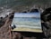 A watercolor painting lies on a rocky shore, depicting a seascape with blue and green hues, small waves, and a city skyline in the distance. The painting is surrounded by art supplies, including brushes and small paint pots.