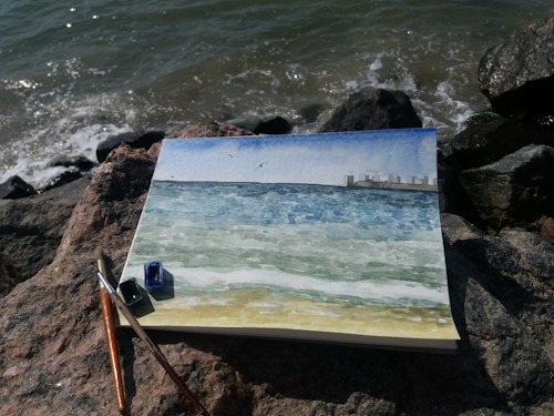 A watercolor painting lies on a rocky shore, depicting a seascape with blue and green hues, small waves, and a city skyline in the distance. The painting is surrounded by art supplies, including brushes and small paint pots.