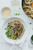 A plate of creamy mushroom risotto is garnished with fresh parsley and shredded cheese, placed on a white tablecloth. There is a bowl of shredded cheese and a glass of white wine nearby. The image conveys a cozy and appetizing setup for a meal.