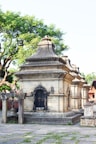A stone temple structure adorned with intricate carvings and a bell hanging from a wooden frame. The building features ornate, arched windows with lattice designs and is surrounded by lush green trees. The area around the temple is paved with stone slabs, some of which have grass growing between them.