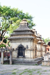 A stone temple structure adorned with intricate carvings and a bell hanging from a wooden frame. The building features ornate, arched windows with lattice designs and is surrounded by lush green trees. The area around the temple is paved with stone slabs, some of which have grass growing between them.